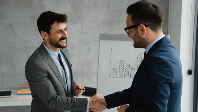 Kunde og rådgiver i håndtrykk foran diagram – et uttrykk for tillit, oppfølging og resultater over tid ved firmareiser fra et reisebyrå for bedrifter.