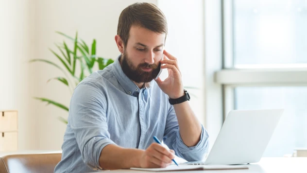 Rådgiver i telefon mens han noterer – et uttrykk for tilgjengelighet og handling når det virkelig haster ved firmareiser fra et reisebyrå for bedrifter.