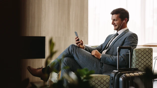 Forretningsreisende i dress sitter avslappet i lounge med koffert og mobil – firmareiser organisert av et reisebyrå for bedrifter.